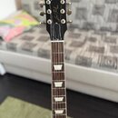 Fotografie zachycuje hmatník a hlavu elektrické kytary s černým povrchem hlavy. Na hlavě je bílý nápis Gibson a pod ním kurzívou Les Paul s doplňkem…