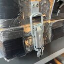 Detailní záběr na kovový zámek kufrů, který je připevněn k povrchu opasanému černou lepicí páskou. Povrch kufrů vykazuje výrazné opotřebení, jsou vid…