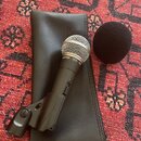 Černý dynamický mikrofon s kovovou síťkou, umístěný na černém koženěném pouzdře. Na těle mikrofonu je viditelný nápis SHURE SM58 a dále v rytině SHUR…