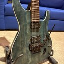 Elektrická kytara s modrozeleným průsvitným povrchem, pod kterým je vidět výrazná struktura dřeva. Nástroj má bílý lem kolem korpusu, dva humbuckery…