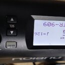 Detailní záběr na ovládací panel černého elektronického modulu. Je vidět displej s nápisy TR-909, DRUM KIT a j=126. Kolem displeje jsou umístěna tlač…