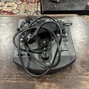 Šedý DJ mixér s ovládacími knoflíky a posuvníky, na kterém leží černý napájecí adaptér a zamotané černé kabely. Na panelu jsou viditelné nápisy CUE,…