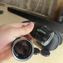 Fotografie zachycuje dvě kovové mřížky mikrofonu v černé barvě. Na jedné z nich je viditelný bílý nápis SHURE. V pozadí je černý textilní obal a dvě…
