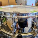 Kovový snare drum s chromovaným povrchem a bílým blátcem. Na obruči je černý nápis SONOR 2.3mm PowerHoop. Na blátci je vidět částečně odlepená nálepk…