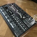 Černý analogový syntetizátor v dřevěném rámu s mnoha ovládacími knoflíky, posuvníky a tlačítky. V pravé části jsou zapojeny bílé patch kabely do sekc…