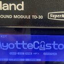 Černý bicí modul s bílými nápisy Roland, DRUM SOUND MODULE TD-30 a SuperNATURAL v oválu. Modrý displej zobrazuje text DRUM KIT a AyotteCustom s ikono…