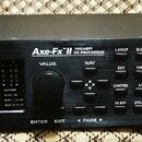 Černý rackový procesor s nápisy FRACTAL AUDIO SYSTEMS a Axe-Fx II PREAMP/ FX PROCESSOR. Na předním panelu je displej, ovládací knoflíky a tlačítka s…