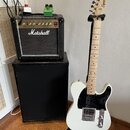 Bílá elektrická kytara značky Squier na černém stojanu. Kytara má černý pickguard a světle dřevěný hmatník. Vedle kytary stojí černý zesilovač Marsha…