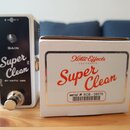 Bílá efekt pedál s černým předním panelem a kovovým spínacím tlačítkem. Na panelu jsou nápisy GAIN, Super Clean a BY xotic usa. Vedle pedálu je bílá…