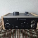 Zadní strana šedého elektronického zařízení s dřevěným rámem. Uprostřed je bílý nápis UNIVERSAL AUDIO s logem UA. Na zadním panelu jsou umístěny vypí…
