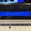 Detailní záběr na modrý displej s bílým textem MCU Extender PRO by LOUD Technologies, Inc. Pod displejem je stříbrný kovový panel s černými poli ozna…