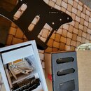 Černý plastový pickguard pro kytaru s výřezy na snímače a potenciometry. Vedle něj je bílá krabička s černým kytarovým mostem a pružinami. V hnědé kr…