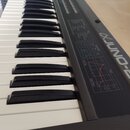 Detailní pohled na klávesnici syntetizátoru s černými a bílými klávesami. Na šedém ovládacím panelu je vidět bílý nápis „JUNO-D“ a logo „Roland“. Pan…