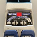 Stříbrný kytarový procesor s modrými spínači, na kterých je bílý nápis DigiTech. Na ovládacím panelu je nápis RP50 MODELING GUITAR PROCESSOR a červen…