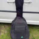 Černý textilní pouzdro (gig bag) na kytaru s přední kapsou. V dolní části kapsy je viditelné tyrkysové trojúhelníkové logo s nečitelným nápisem uvnit…