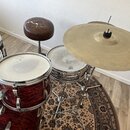 Sada červených bubnů s tmavým pruhovaným vzorem a stříbrným kováním. Na jednom z kotlů je viditelné logo SONOR. Součástí je mosazný činel na chromova…