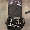 Černý přepravní kufr s rukojetí, ve kterém jsou uloženy části kovového bubnového pedálu stříbrné barvy. V horní části je vidět krabice s obrázkem ste…