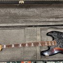 Elektrická basová kytara v barvě sunburst s černým perleťovým pickguardem, umístěná v otevřeném tvrdém kufru. Na hlavě nástroje je viditelné logo Fen…