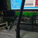 Černý ramenní mikrofonní stojan připevněný k šedému stolu. V pozadí je vidět počítačový monitor značky msi, klávesnice a audio rozhraní s nápisy behr…