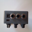 Fotografie zachycuje zadní stranu šedého elektronického zařízení s plastovým povrchem. Jsou zde vidět tři 6,3mm jack konektory, jeden konektor pro na…