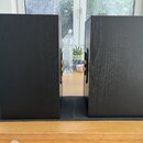Dva černé reproduktory s povrchem imitujícím strukturu dřeva. Stojí vedle sebe na černých podložkách na dřevěném stole. Na reproduktorech nejsou vidi…