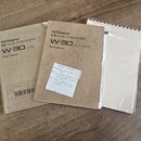 Fotografie zobrazuje tři dokumenty k hudebnímu nástroji položené na dřevěném povrchu. Jsou zde dva tištěné manuály s nápisy „Roland MIDI MUSIC WORKST…