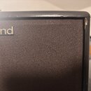 Detailní záběr na horní roh černého komba. Na přední textilní mřížce je vidět část loga Roland. Horní hrana šasi vykazuje drobné oděrky a v pravém ro…