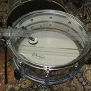 Fotografie zachycuje snare drum s chromovaným obručováním a bílým koženem. Na koženě je viditelné logo s nápisem „Ludwig“. Přes buben je přeložen kov…