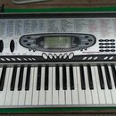 Elektronický klavír značky CASIO v bílo-šedém provedení s černými klávesami. Na horní části je rozsáhlý seznam funkcí a tónů v sekcích 137 TONES, 100…