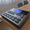 Šedý elektronický bicí modul s modrým displejem a velkým ovládacím knoflíkem. Na horní části je viditelné logo Roland. Zařízení obsahuje množství gum…