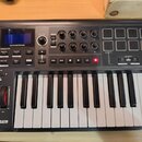 Černý MIDI kontrolér s klávesnicí a červenými bočními panely. Na zařízení je vidět logo novation, nápis IMPULSE 25 a ovládací prvky jako pady, knoflí…