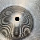 Detailní pohled na střed kovového činele stříbrné barvy. Povrch je voorzien výraznými koncentrickými drážkami. V levém horním rohu je viditelný fragm…