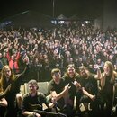 Fotografie zachycuje skupinu šesti hudebníků na pódiu před velkým množstvím lidí. V popředí je muž s kytarou, u kterého není značka nástroje čitelná.…