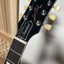 Černá hlava elektrické kytary s bílými nápisy Gibson, Les Paul MODEL a Standard. Nástroj má šest ladicích kol v krémové barvě a hnědý hmatník s trape…