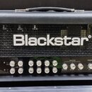 Černý kytarový zesilovač s kovovou mřížkou, na které je bílý nápis Blackstar. Přední panel obsahuje řadu ovládacích knoflíků a přepínačů s popisky ja…