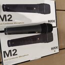 Tři černé dynamické mikrofony s mřížkou, položené na dvou krabicích. Na tělech mikrofonů je nápis RODE M2 a na krabicích texty M2, Live Condenser Mic…