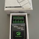 Efektový pedál v bílém kartonovém boxu s nápisem PEDAL EFFECT a nákresem komba. Pedál je zelený a černý s nápisy OUTPUT, INPUT, BASS LIMITER a BLM-I.…