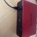 Červený audio rozhraní s kovovým povrchem a černým zadním panelem. Na horní a boční straně je nápis Focusrite. Zadní panel obsahuje USB konektor s př…