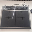 Šedý MIDI kontrolér s osmi černými gumovými pady. V levém horním rohu je bílé logo ALESIS a v pravém horním rohu nápisy ControlPad, USB/MIDI Percussi…