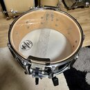 Fotografie zachycuje pohled z horní strany do otevřeného bubnu. Viditelný je světlý dřevěný korpus, spodní bílá blana s nápisem EVANS RESONANT SNARE…