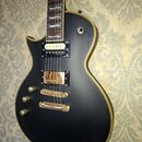 Černá elektrická kytara s jednodrážkovým tělem a zlatými detaily na hranách. Nástroj má dva humbuckery, přičemž horní je bílý a dolní zlatý. Na černé…