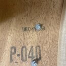 Detail dřevěného povrchu s dvěma kovovými šrouby. Na dřevě jsou černým tiskem nápisy „DEO 11 58“ a pod ním větší nápis „P-040“. Povrch vykazuje přiro…