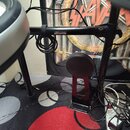 Černý kovový stojan na elektronické bicí s připevněným malým černým sedákem. Na vodorovné tyči je viditelný bílý nápis YAMAHA. Kolem konstrukce jsou…
