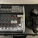 Míchadací pult značky behringer, model XENYX QX1202USB, umístěný v černém přepravním kufru. Zařízení má stříbrno-černý povrch s řadou knofíků, posuvn…
