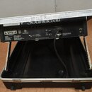 Zadní strana elektronického zařízení v černé barvě, umístěná v otevřeném transportním kufru s hliníkovými hranami. Na horním bílém panelu je částečně…