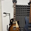 Elektrická kytara s tónem sunburst v hnědých odstínech a krčkem z tmavého dřeva. Na hlavě je bílý nápis Gibson a pod ním stylizovaný nápis Les Paul.…