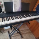 Černé digitální piano umístěné na černém X-kovovém stojanu. Na přístroji je viditelné logo YAMAHA a na přiloženém manuálu nápisy DIGITAL PIANO P-115.…