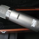 Kondenzátorový mikrofon stříbrné barvy s kovovou síťkou v dřevěném kufru s černým polstrováním. Na těle je černý nápis WARM AUDIO a bílá nálepka s te…