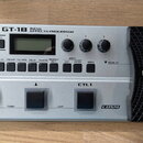 Stříbrný basový efektový procesor s černým výrazným pedálem vpravo. Na horní části je nápis BOSS GT-1B BASS EFFECTS PROCESSOR, tři ovládací knoflíky…