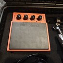 Oranžový elektronický perkusní pad s černou gumovou údernou plochou, na které je bílý nápis Roland. V horní části jsou čtyři černé otočné ovladače a…